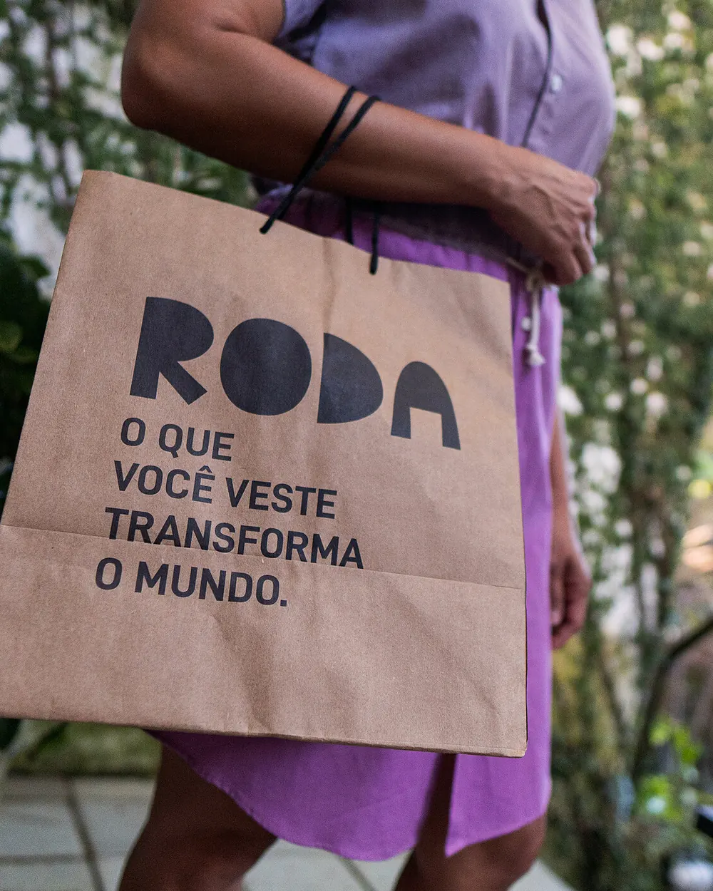 Sacola de papel da RODA com a frase “o que você veste transforma o mundo”, carregada por uma pessoa de vestido lilás.
