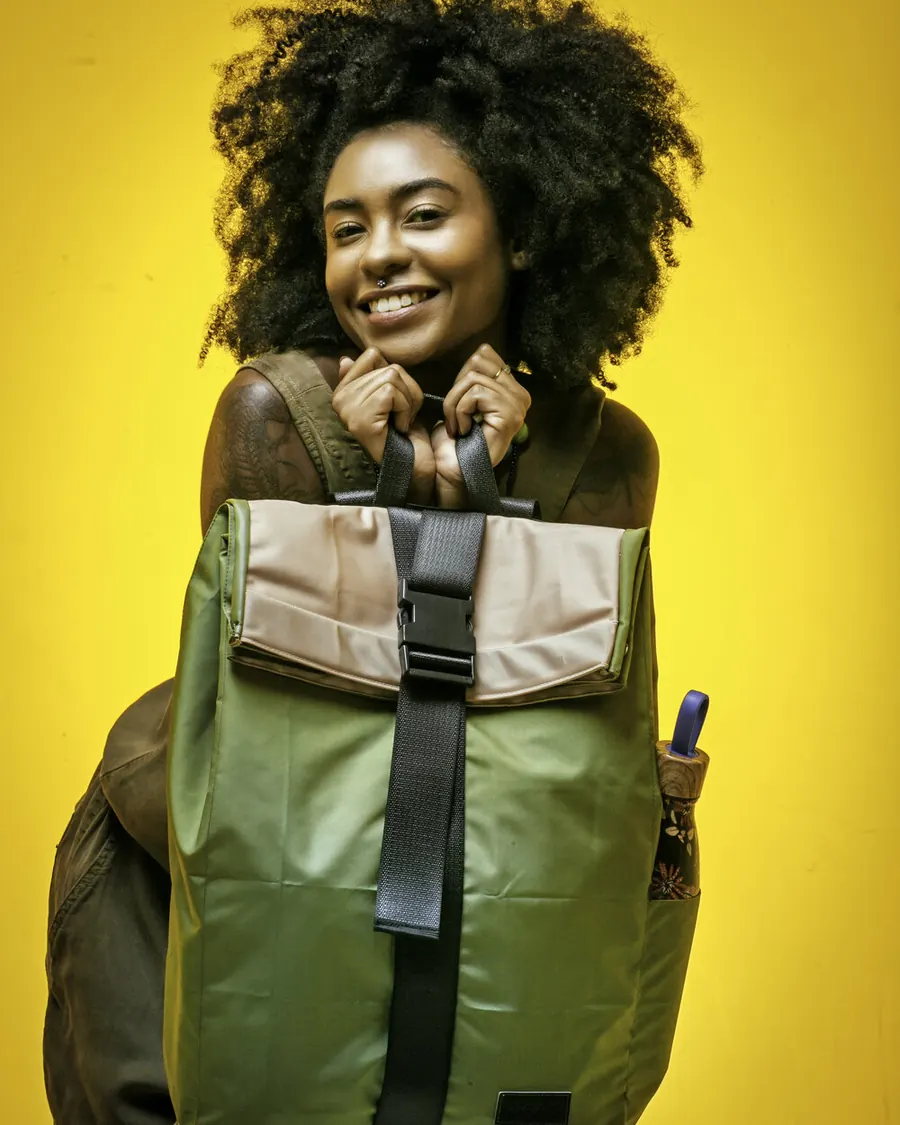 Pessoa sorrindo com mochila verde da RODA, feita de airbag, de fecho preto, em fundo amarelo.